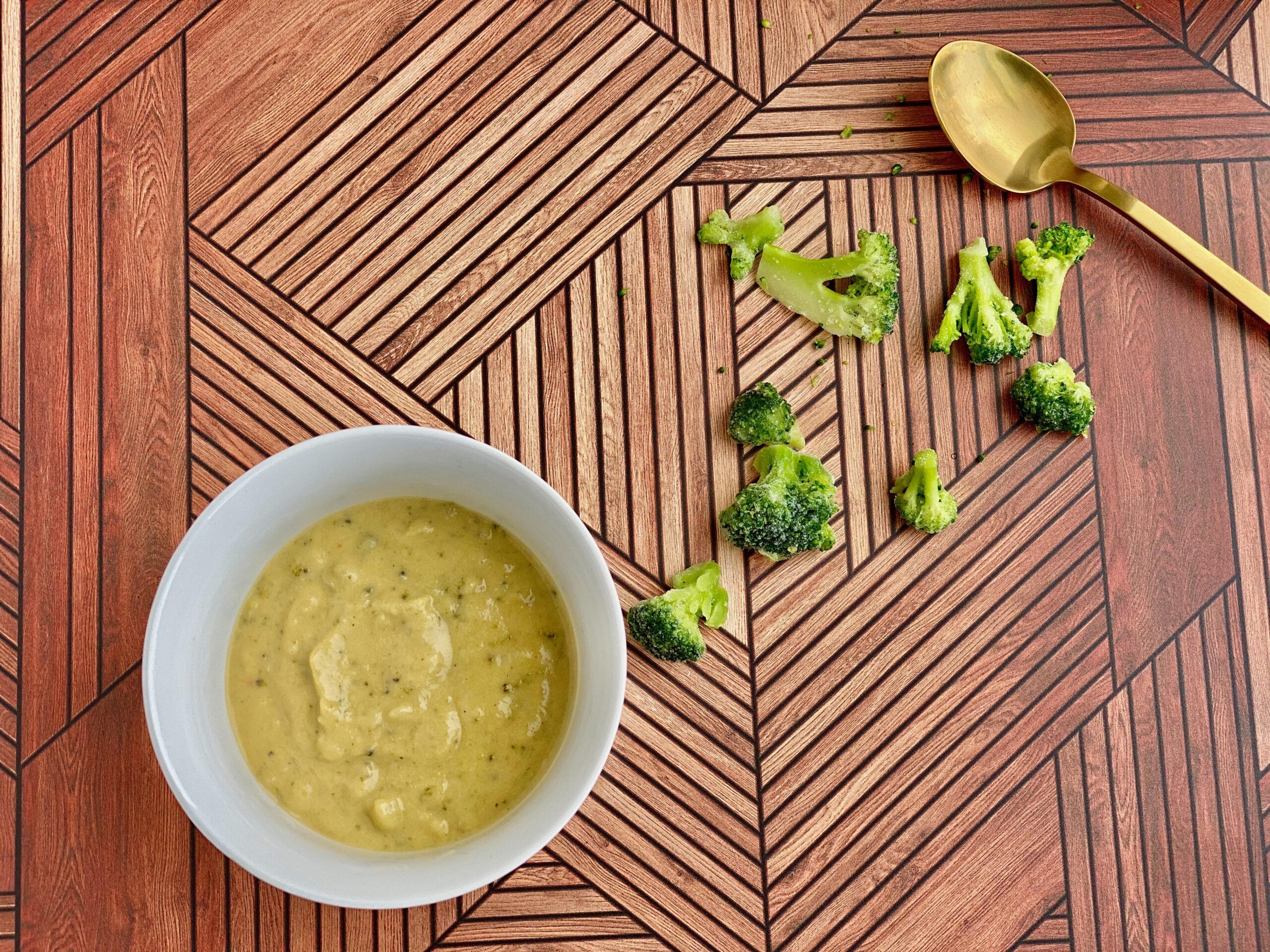 Easy Broccoli Cheddar Soup Image of a bowl of soup with pieces of broccoli and a spoon in the background