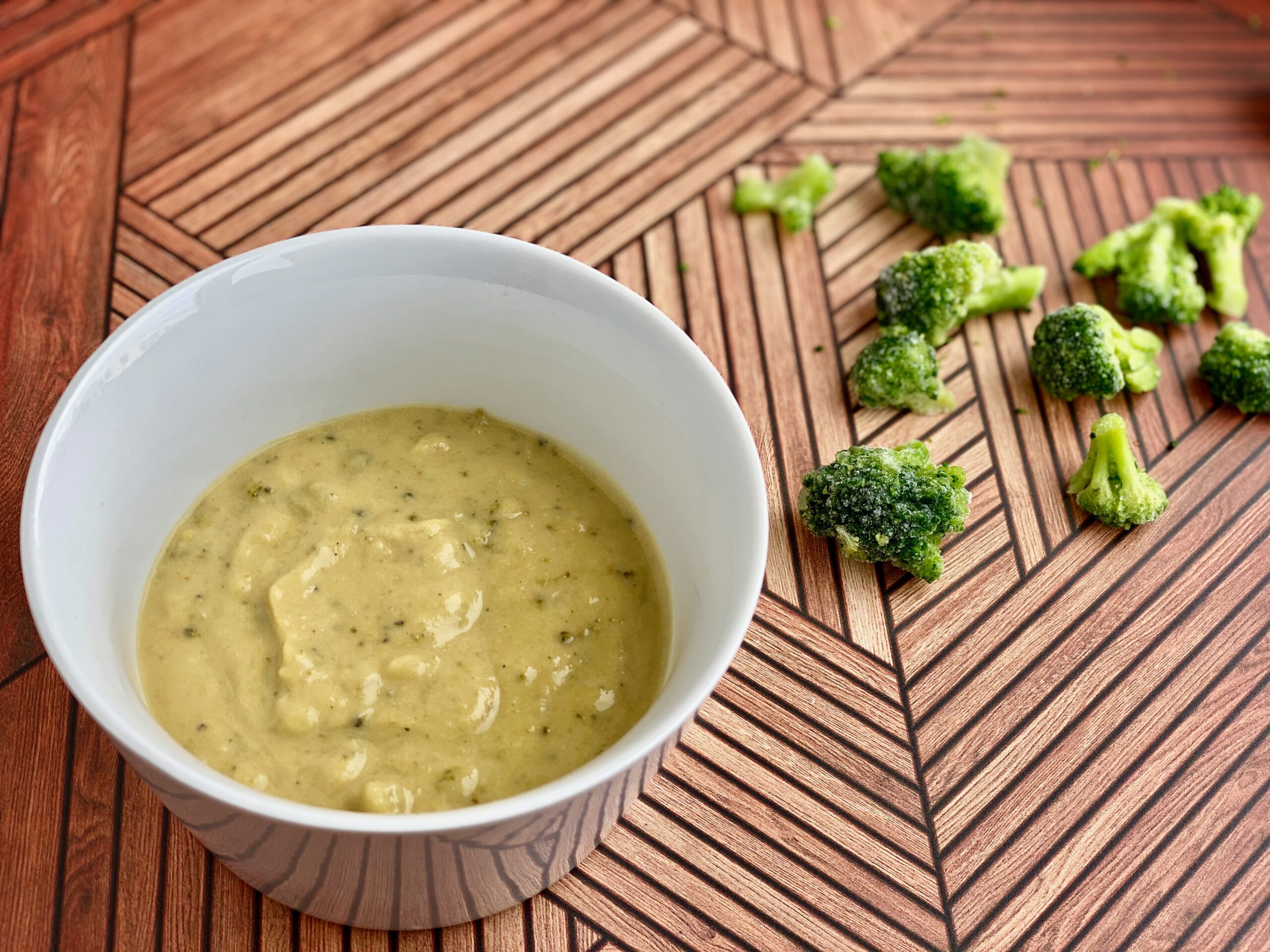 Easy Broccoli Cheddar Soup Image of a bowl of soup with pieces of broccoli in the background