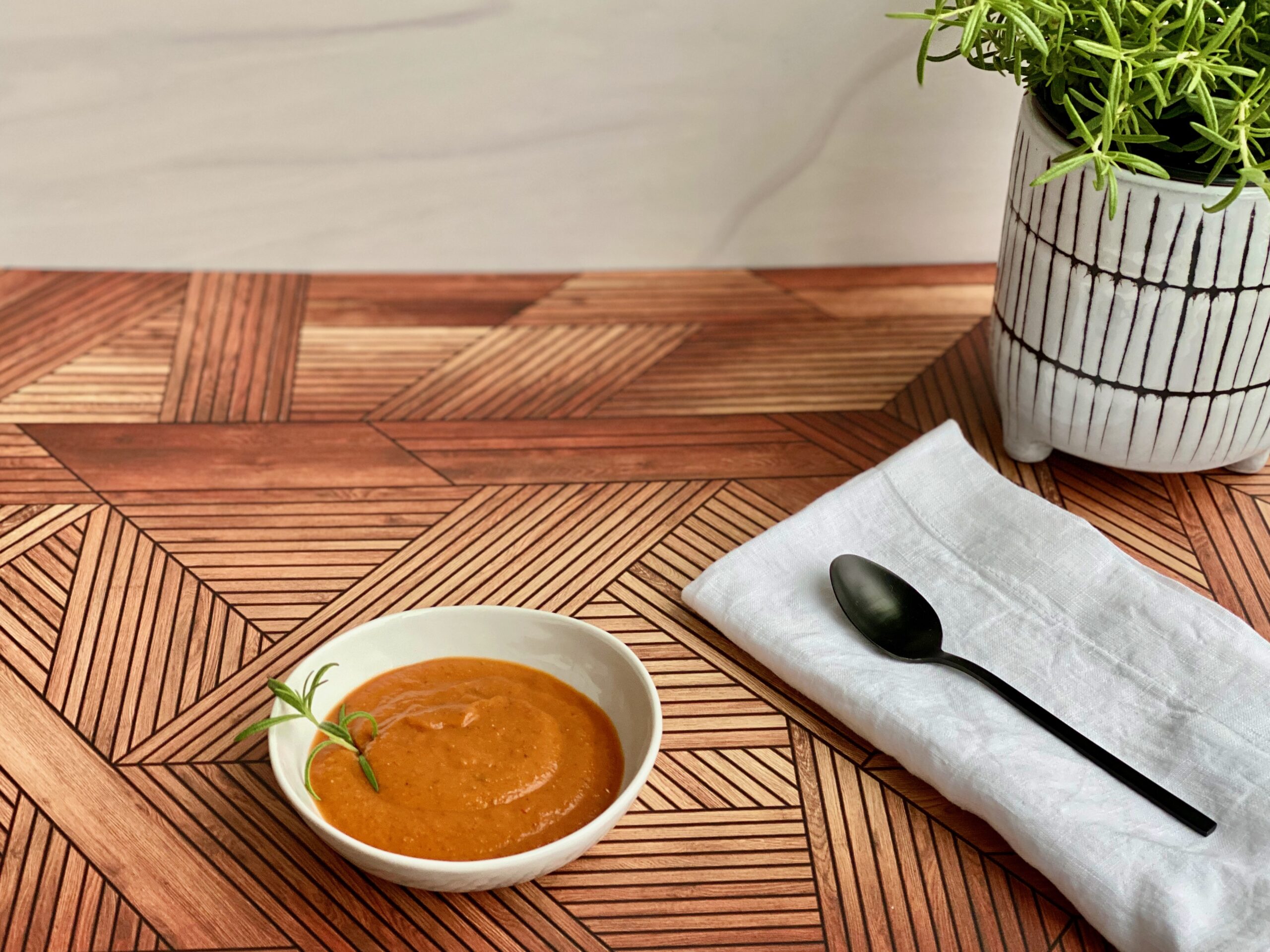 Easy Roasted Tomato Vegetable Soup An image of a bowl of tomato soup with a rosemary plant in the background